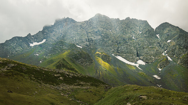 Jagged Caucasus peaks rise above green alpine slopes and lingering snow near the Abudelauri Lakes in Georgia, conveying a dramatic, wild and mysterious high-mountain atmosphere