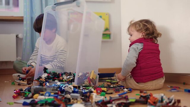 Children building separately with interlocking blocks on a bedroom floor, seated on opposite sides of an upright transparent storage bin surrounded by loose pieces