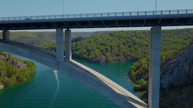Aerial drone view of Krka Bridge in Croatia, flying smoothly through a concrete pillar as cars cross the bridge above turquoise river waters surrounded by rocky, forested landscape.
