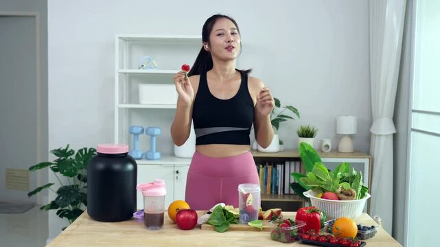 Smiling Asian fitness influencer recording a health podcast, scooping pink protein powder into a shaker bottle. Woman vlogger creating sports nutrition and healthy diet content with fresh food.