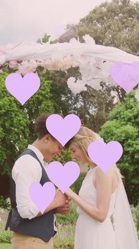 Animation of lilac hearts and happy diverse couple holding hands under wedding arch in garden