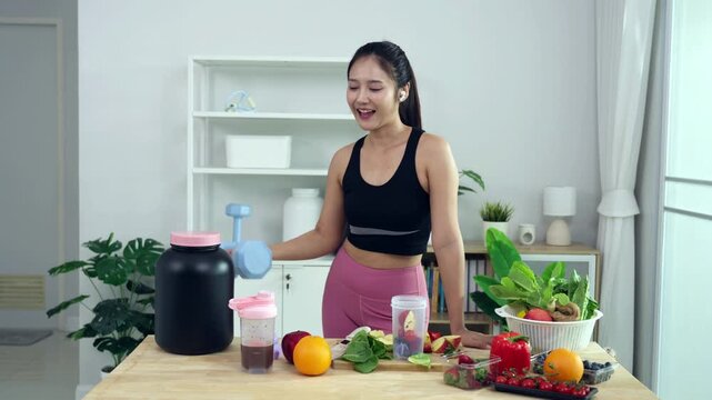 Smiling Asian fitness influencer recording a health podcast, scooping pink protein powder into a shaker bottle. Woman vlogger creating sports nutrition and healthy diet content with fresh food.