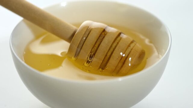 Wooden honey dipper dipping into golden honey in white ceramic cup, sweet natural food close up