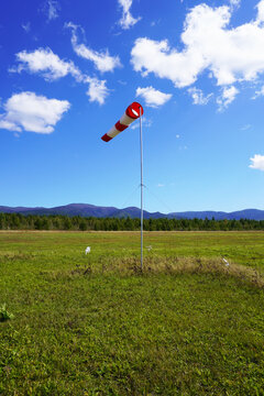 Cone wind indicator. Windsock on pole over grassy field. Red windsock against blue sky, green grass, distant trees. Open rural area.