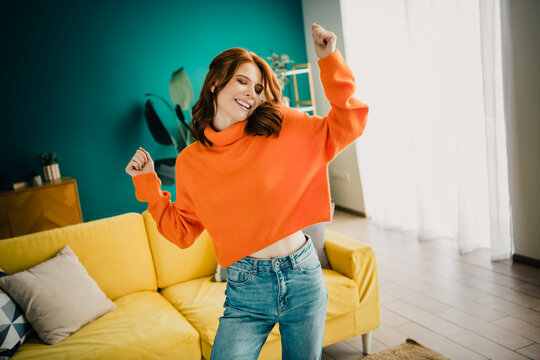 Happy young woman dancing in living room wearing bright orange sweater and jeans, enjoying music and relaxed home lifestyle