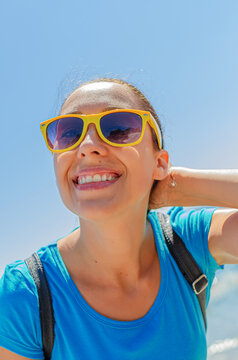 A happy young woman in yellow sunglasses smiles under a clear blue sky during a sunny summer day.