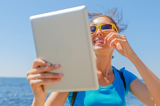 A smiling young woman wearing sunglasses holds a digital tablet near the ocean on a bright sunny day.