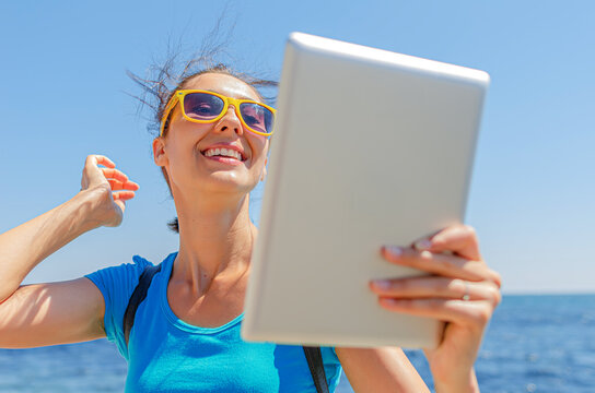A happy young woman wearing sunglasses smiles while taking a selfie with a digital tablet outdoors near the sea on a sunny day.