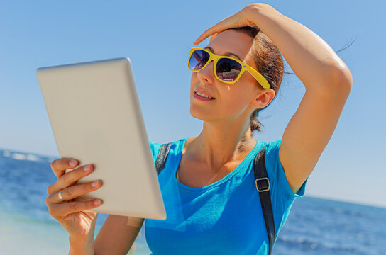 A woman in sunglasses holds a digital tablet while relaxing near the bright blue sea on a sunny day.