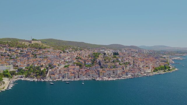 Aerial drone view of &Scaron;ibenik, Croatia, with a smooth orbital camera movement revealing the historic waterfront, dense red rooftops, medieval architecture, blue Adriatic Sea, boats and hills.