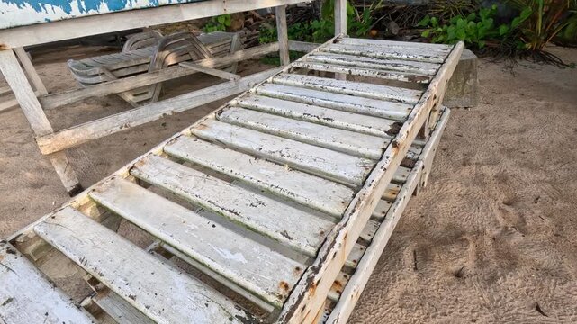 Old unpainted wooden sun lounger stands on wet sand surrounded by old loungers, tables, and chairs at a coastal beach area during daytime.