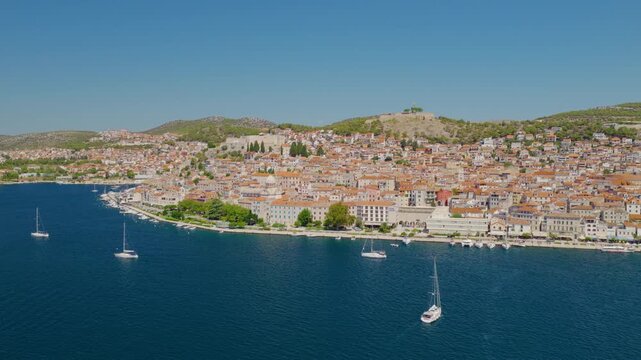 Aerial drone view of &Scaron;ibenik, Croatia, with a smooth orbital camera movement revealing the historic waterfront, dense red rooftops, medieval architecture, blue Adriatic Sea, boats and hills.