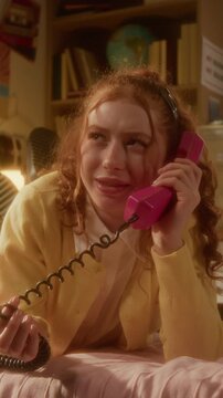 Vertical video of Caucasian teenager lying on bed, talking on retro pink landline telephone and twirling cord. Ideal for nostalgic youth lifestyle commercials or retro technology marketing