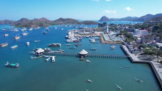 Aerial drone footage approaching the marina and harbor waterfront of Labuan Bajo in Flores, Indonesia. The scene features a long pier, numerous tour boats and speedboats, and marina facilities