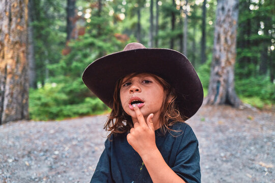 Boy in a cowboy hat touching his lip at a forest campsite