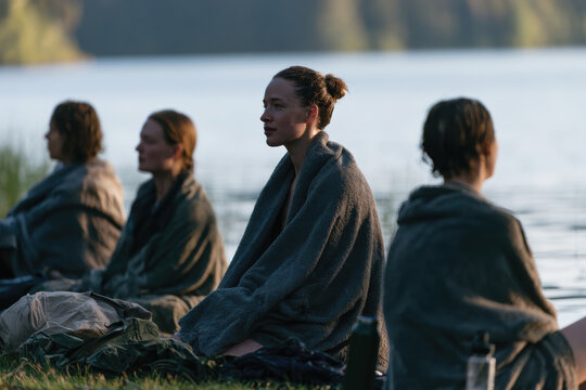 A serene gathering of individuals wrapped in blankets by a reflective lake, immersed in thoughts, surrounded by calm water and nature, highlighting introspection and unity.