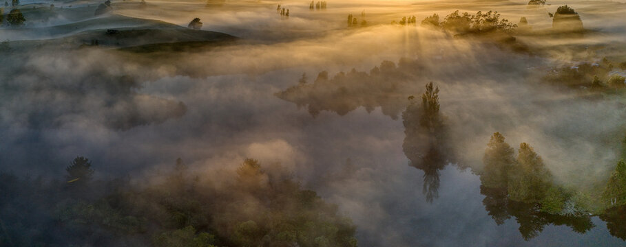 Aerial view of Lake Ratapiko shrouded in morning mist with golden sunlight filtering through trees and rolling hills in Ratapiko, Taranaki Region, New Zealand.