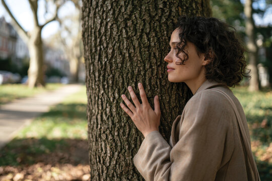 A woman with curly hair gently embraces a tree in a peaceful park setting, highlighting a connection with nature and a moment of introspection amidst urban surroundings.