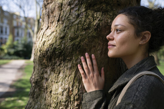 A woman with freckles and curly hair stands pensively by a tree in a sunlit park, symbolizing harmony with nature and moments of personal contemplation.