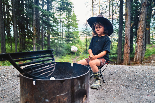 Boy in a cowboy hat holding a roasted marshmallow on a stick by a fire pit
