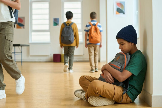 Sad bullied boy sitting alone on the floor in a school hallway with backpack
