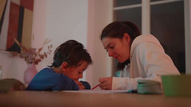 School-age boy concentrates on completing homework beside his mother at the dining table at home, developing independence, diligence, knowledge and academic confidence