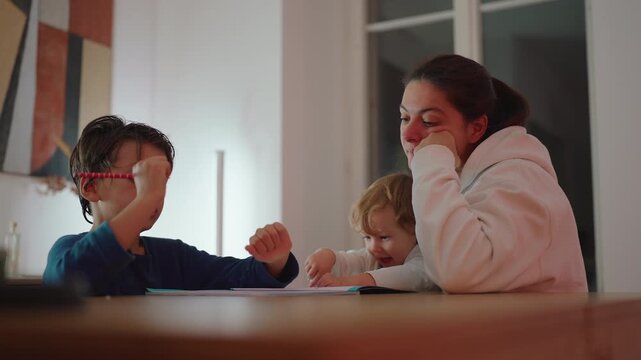 Parent supports young son writing on a worksheet at home while toddler sibling joins them at the dining table, expressing family togetherness, care and shared learning