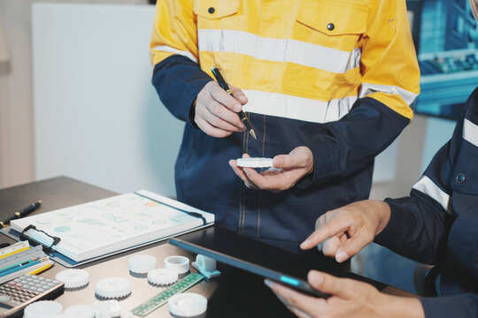Two Asian male industrial engineers, wearing helmets, safety glasses, and uniforms, are working together in a lab. They are examining and analyzing white mechanical gear parts printed with a 3D printe