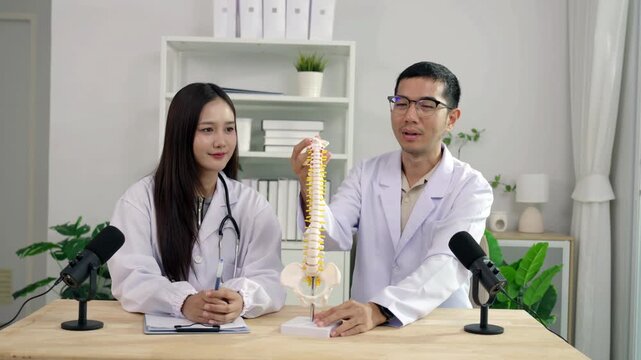Asian female doctor pointing at a spine model explaining orthopedics to a male doctor during a medical podcast interview. Recording a healthcare audio broadcast in a modern clinic studio.