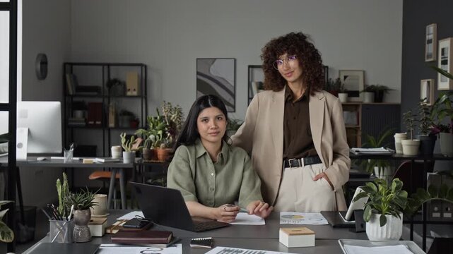Portrait of young adult Latin woman sitting at desk and young adult Caucasian colleague standing in office, smiling at camera. Video represents female leadership and workplace diversity