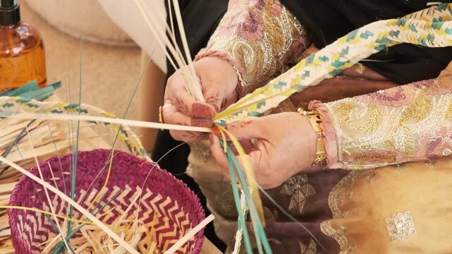 Hands weave pale palm fronds into tight bands, shaping traditional Al Khoos work with steady fingers. Rich dress fabric and dyed baskets add Bedouin inspired cultural detail beside woven basket.