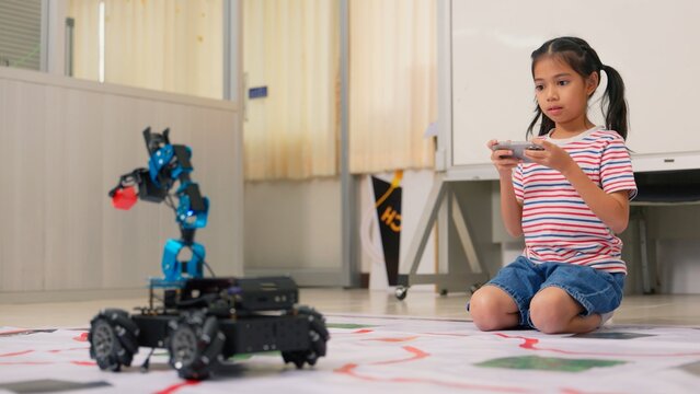 Asian girl student controlling robotic arm car with remote controller in STEM classroom. Science technology engineering mathematics education, creative robotics coding and school study concept.