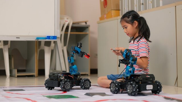 Asian girl student controlling robotic arm car with remote controller in STEM classroom. Science technology engineering mathematics education, creative robotics coding and school study concept.