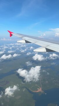 aerial view from an AirAsia flight window over Singapore, showing the plane's red-tipped wing above clouds, with cargo ships dotting the blue sea and coastline below