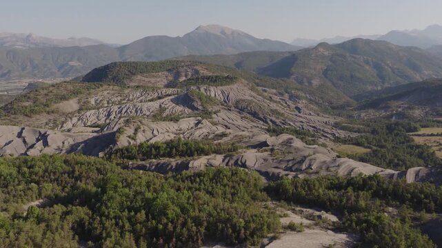 Aerial panorama of heavily eroded gray marl hills with deep gullies sparse pines and forested mountain backdrop