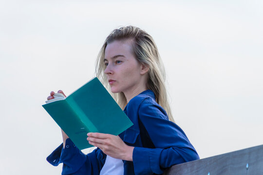 Young blonde woman holding and reading book outdoors against clear sky background