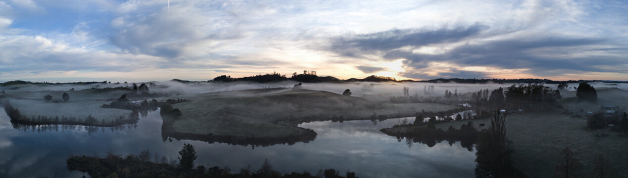 Aerial view of Lake Ratapiko surrounded by rolling hills and dense morning mist under a soft sunrise sky in Ratapiko, Taranaki Region, New Zealand.