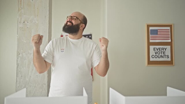 Man wearing white tshirt and ivoted sticker raises clenched fists and looks upward at an american flag and every vote counts poster in a voting booth inside a building; civic pride.