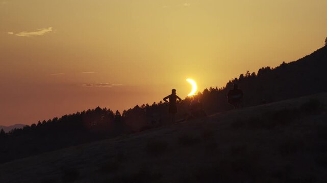 Silhouettes of people watching partial solar eclipse on hillside with golden sky and forested ridge
