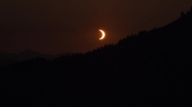 Close view of partial solar eclipse crescent sun glowing orange above dark forested mountain silhouette