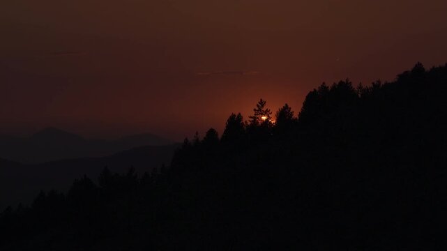 Bright partial solar eclipse behind pine trees on dark ridge with layered mountains and orange sky