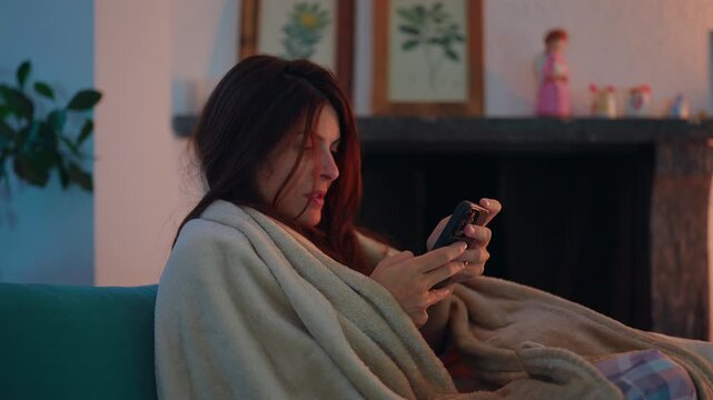 Young woman browses smartphone under a blanket on living room sofa beside fireplace, enjoying cozy leisure, digital connection, personal downtime and comfort at home