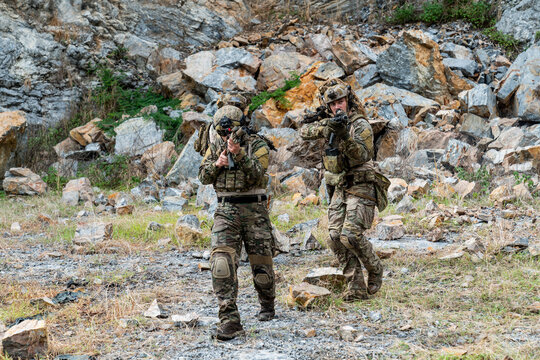 Military personnel engage in tactical training in a rocky outdoor area during daylight