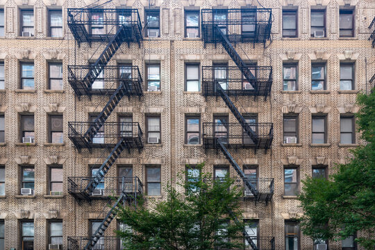 A close-up view of the symmetrical brick facade of a traditional New York residential building, complete with a network of external fire escapes in the Chelsea neighborhood of Manhattan.