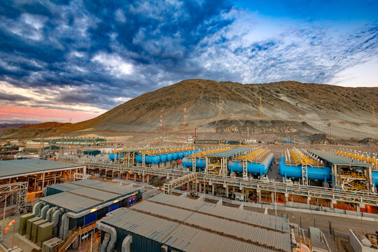 High angle view of a vast reverse osmosis plant along the coast of the Atacama Desert in Chile with large blue tanks and industrial infrastructure set against a bleak mountain landscape.