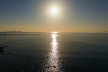 Obraz premium Golden evening sunlight reflecting across calm ocean surface at sunset, gentle swell on sea with distant headland on horizon under clear dusk sky