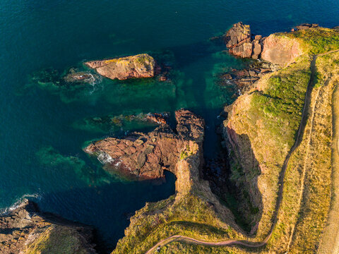 Rocky cliffs and red sandstone sea stacks on Pembrokeshire coastline, Wales, warm evening sunlight illuminating grassy headland and turquoise water, aerial view