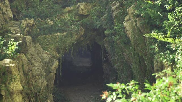 Shadowed tunnel opening at rock face, narrow path leads into dim interior flanked by moss and ivy, rugged stone textures and layered cliffs evoke exploration