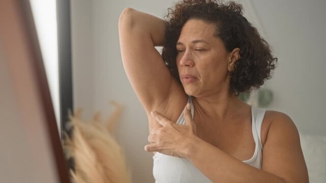 Woman lifts arm and checks armpit skin at mirror in building, touching underarm to inspect for irritation; self care concern.