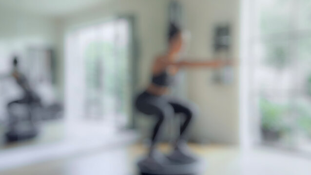 A blurred image of a woman performing squats on a fitness platform in a bright home gym with mirrors and greenery visible outside.